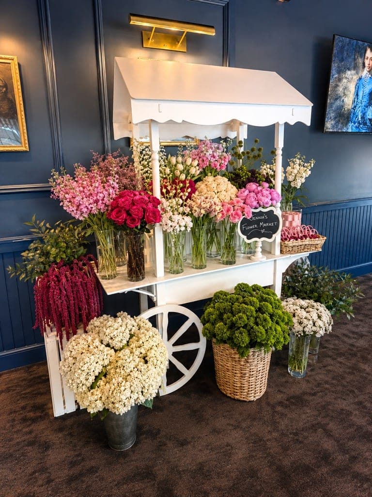 A white flower cart filled with roses, hydrangeas and peonies for a bridal shower flower bar