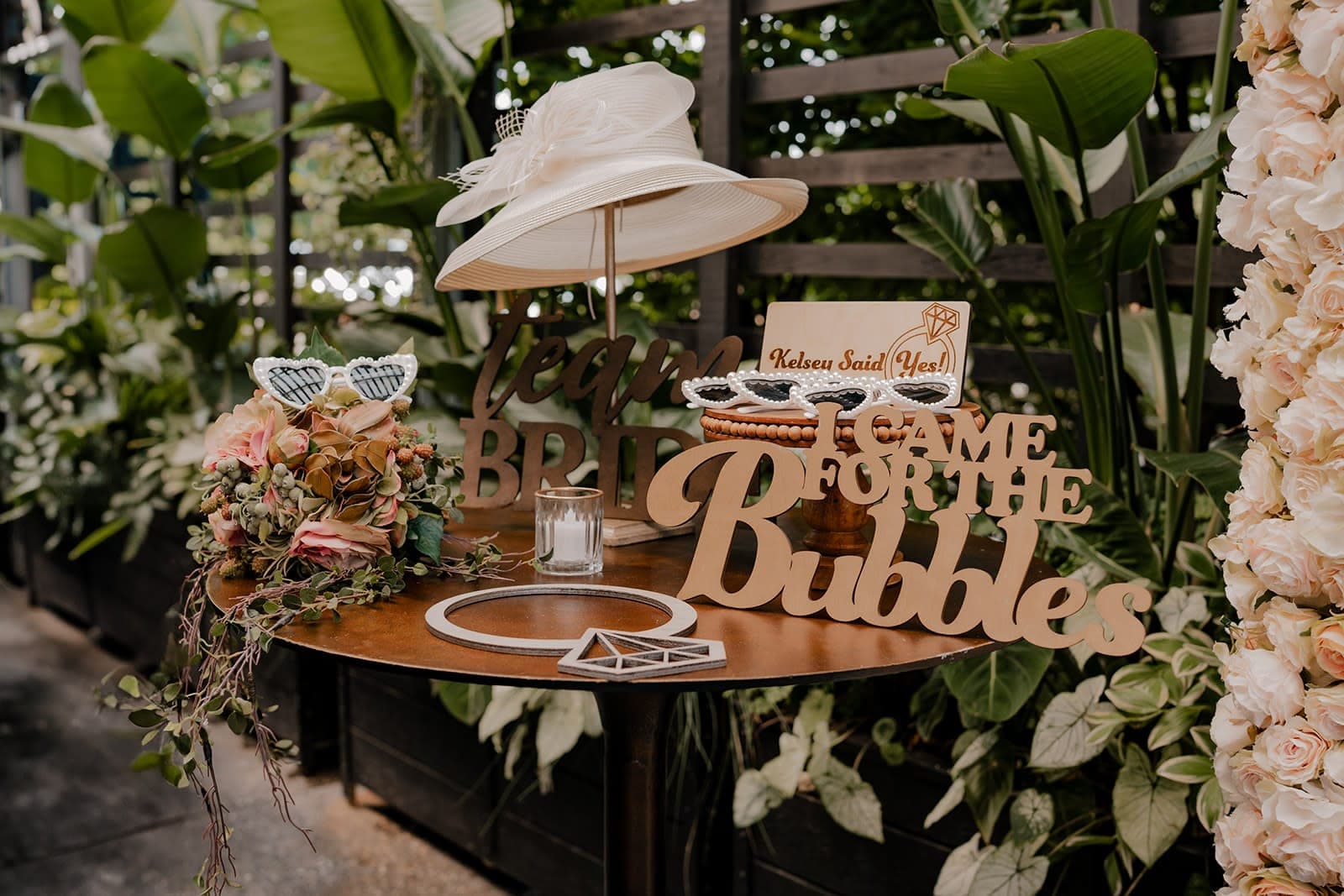 A photo booth props table with a white derby hat, heart sunglasses and a Team Bride sign